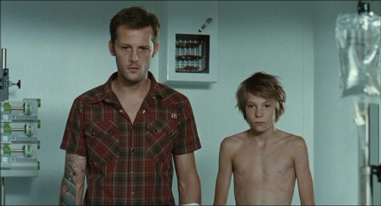 Even in the stark, clinical environment of a hospital, Louis (Steve Le Roi) remains inexplicably bare-chested beside his older brother Julien (Nicolas Duvauchelle)—a deliberate visual choice that trades strict narrative realism for a constant, glaring reminder of his unprotected vulnerability.