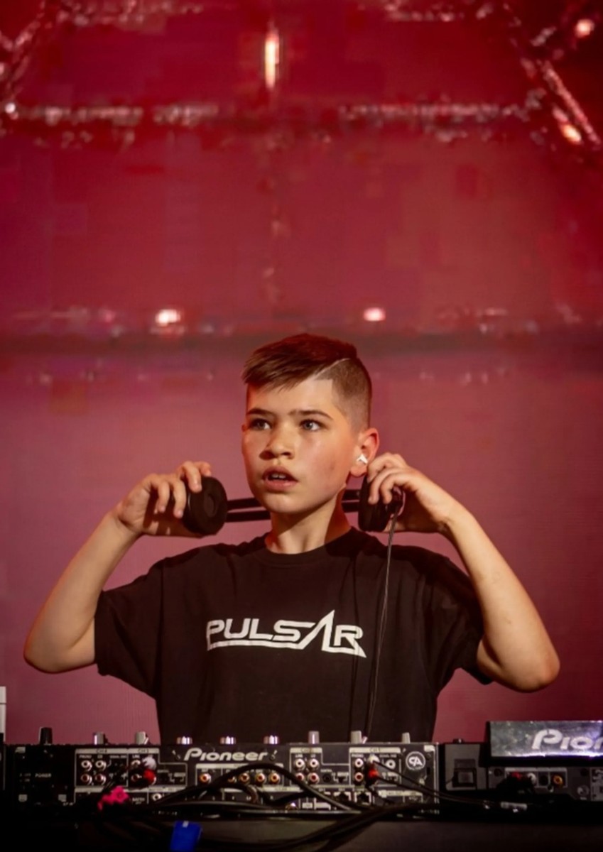 Pulsar performing live behind Pioneer decks under red stage light, headphones held behind his neck